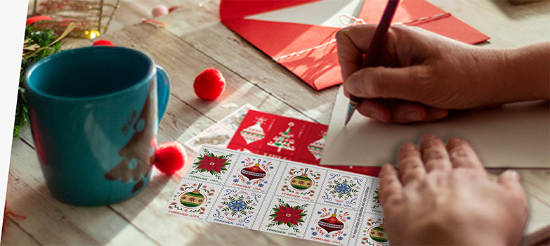 A person addressing a letter with First-Class Mail Forever® holiday stamps on the table.